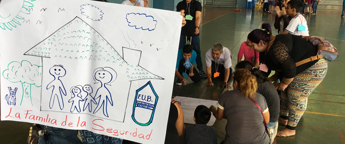 Familias desarrollando dinmica de grupo y un cartel en primer plano con una casa dibujada, dentro de esta una familia y bajo la misma el texto  `La familia de la seguridad`